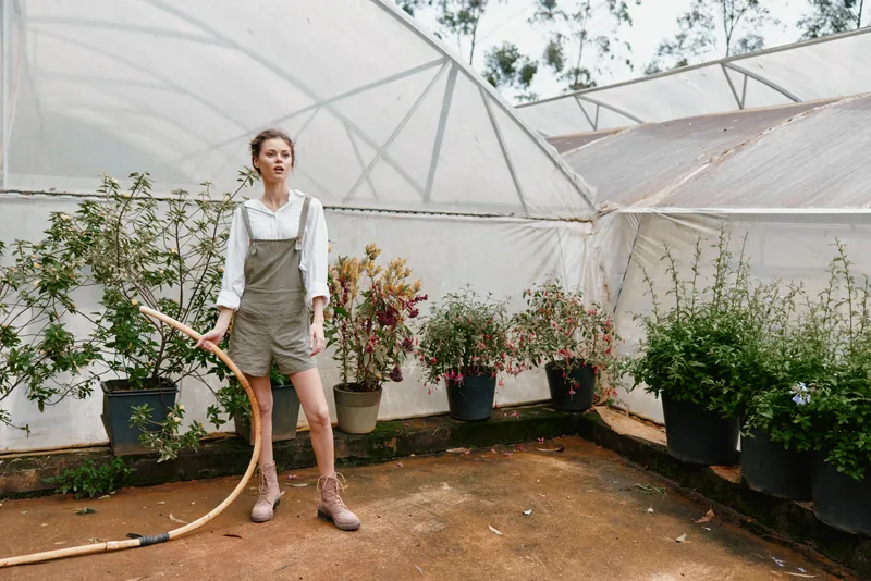 Young woman gardening in a greenhouse, wearing stylish overalls, standing with a hose, surrounded by lush green plants, showcasing a refreshing and vibrant atmosphere
