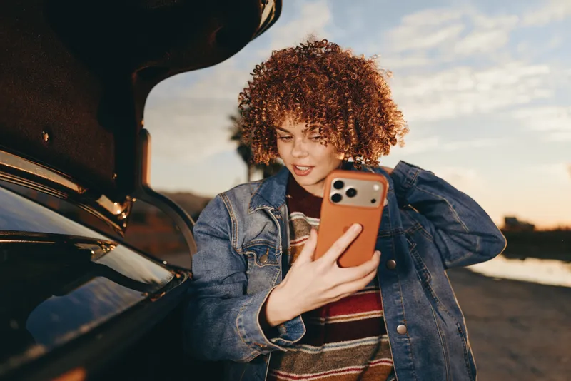 Woman lifestyle outdoors beside a car at sunset, smiling and using smartphone, denim jacket, curly hair, casual warm light, soft evening breeze, near parking lot, natural colors today