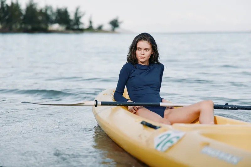 A focused woman sits in a yellow kayak on calm water, wearing a navy long sleeve top, holding a paddle, ready for a peaceful paddle session on open water with soft light and tranquil surroundings.