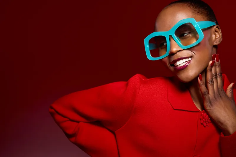 Stylish African woman with dark skin wearing oversized blue glasses and red sweater smiles confidently against a red background. Concept of fashion, beauty, and modern style.