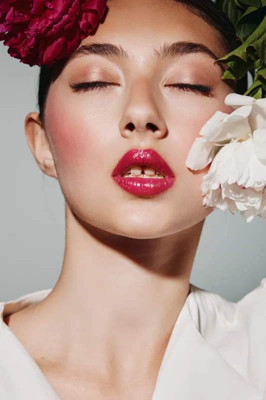 Close-up portrait of a woman with glossy red lips and soft rosy makeup. She poses with closed eyes and flowers near face in a high-fashion beauty editorial style.