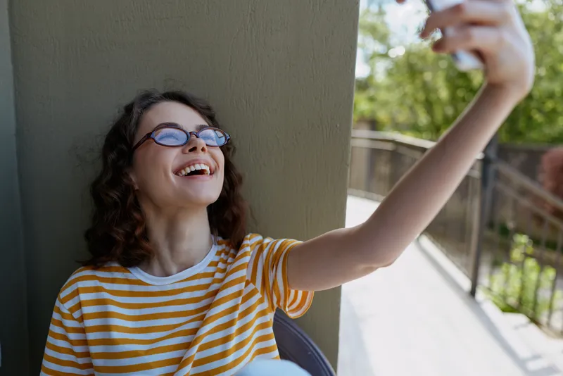 woman lifestyle outdoor selfie smiling on balcony wearing striped yellow shirt casual sunlit moment relaxed pose natural colors greenery background bright day joyful mood