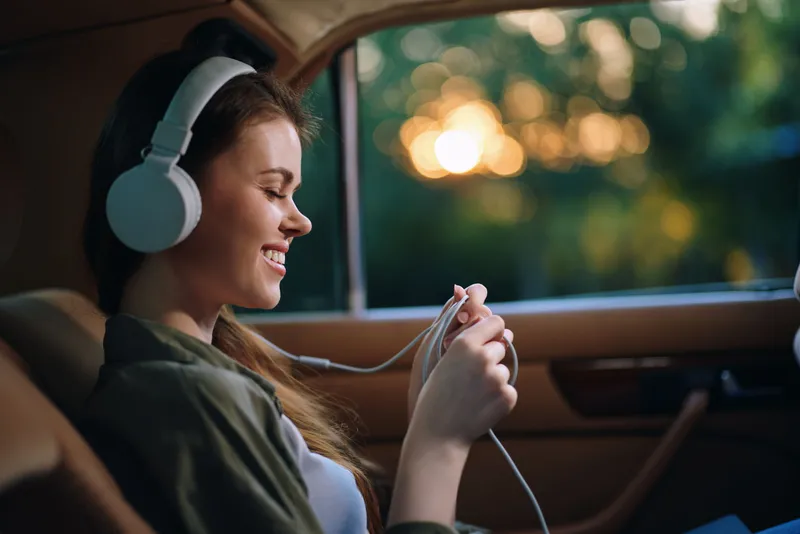 Young woman enjoying music in a car with headphones, smiling, vibrant sunset background, relaxed mood, emphasizing leisure and comfort.