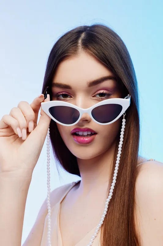 Young woman with long hair wearing white sunglasses and pink makeup poses against a blue background. Concept of woman beauty fashion and modern style.