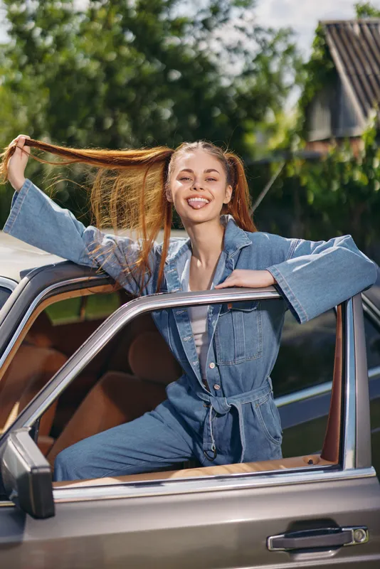 Smiling young woman in denim jumpsuit, playfully posing with long hair outside vintage car during sunny day in green nature.