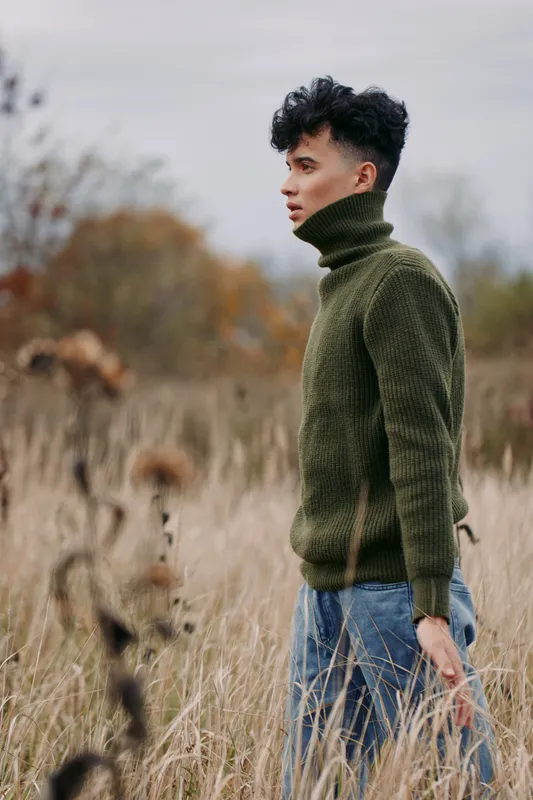 A candid portrait of a lone model standing in tall grass, wearing a forest green knit turtleneck, conveying authenticity and quiet realism in natural light.