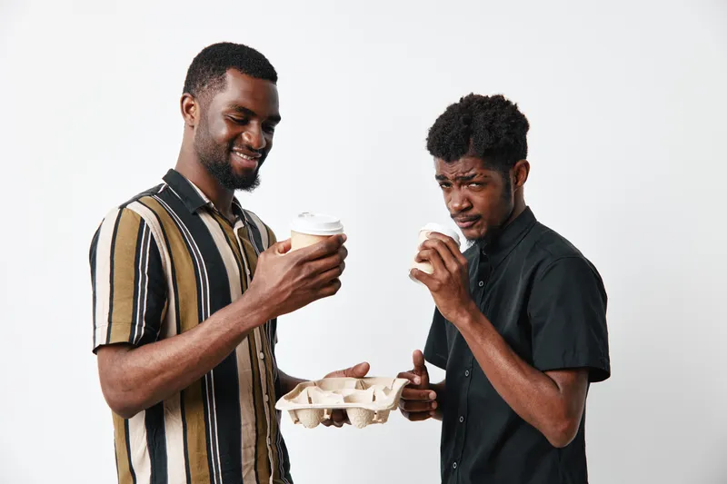 Two african american men with dark skin holding coffee cups and a cardboard tray smiling and drinking together on white background in casual clothing and relaxed mood.