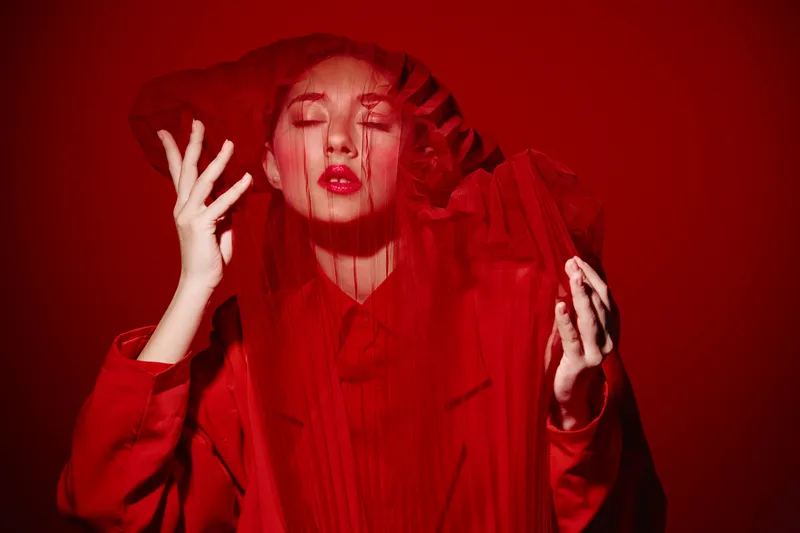 Woman in dramatic red outfit with sheer fabric covering her face poses artistically in studio with red background. Beauty fashion concept with expressive makeup tones and elegant styling.