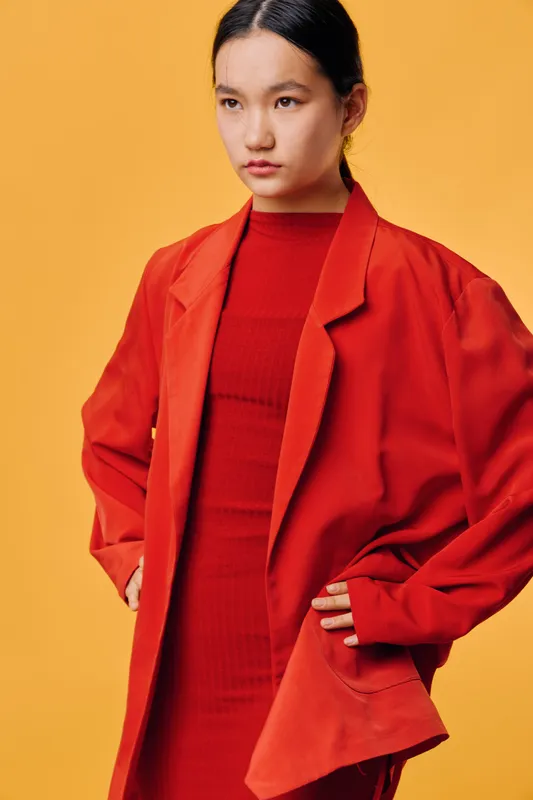 Woman wearing red dress and oversized jacket posing against orange background with confident expression and hands on hips. Fashion, style, portrait and vibrant color contrast.
