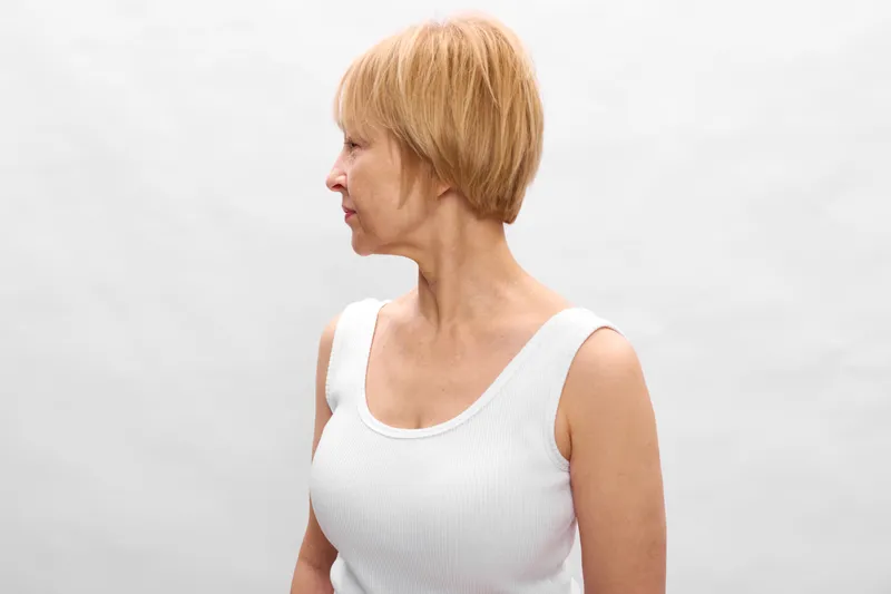 Calm woman, mature profile, short blonde hair, white tank top, bright studio background, concept of aging, health, wellness, peaceful mood, studio portrait, relaxed posture, calmly