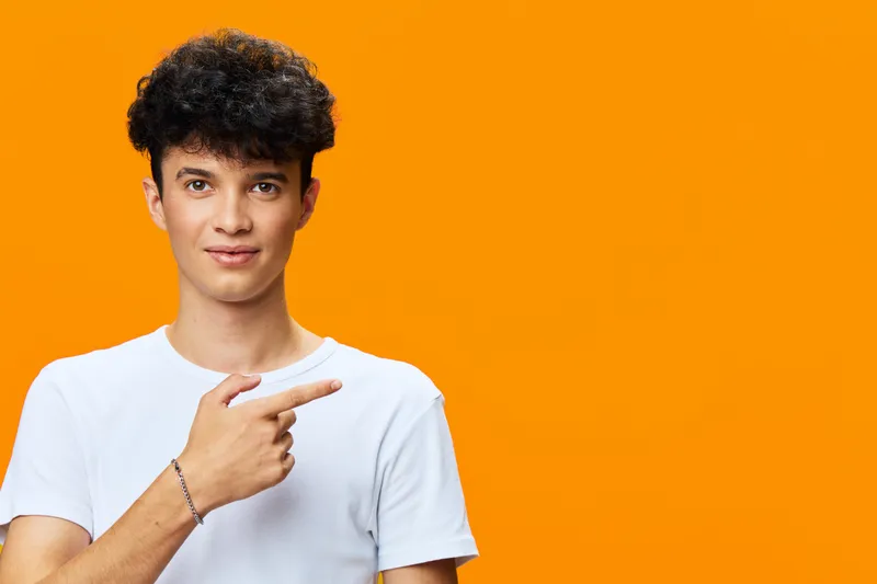 Young man in white t-shirt pointing sideways with finger, curly hair, friendly expression on bright orange background, casual style portrait.