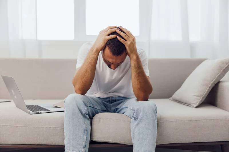 Stressed man sitting on a couch, holding his head in despair while working on a laptop. The scene evokes feelings of anxiety and overwhelming challenges in a modern setting.