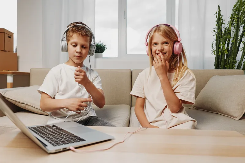 Two children with headphones enjoy music and play at home using a laptop. Cozy living room setting with natural light and casual clothes for entertainment and fun.