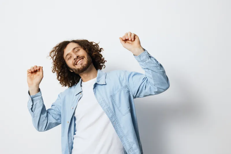 Happy young man with curly hair dancing joyfully in a light blue shirt, depicting a vibrant and energetic lifestyle against a minimalist white background
