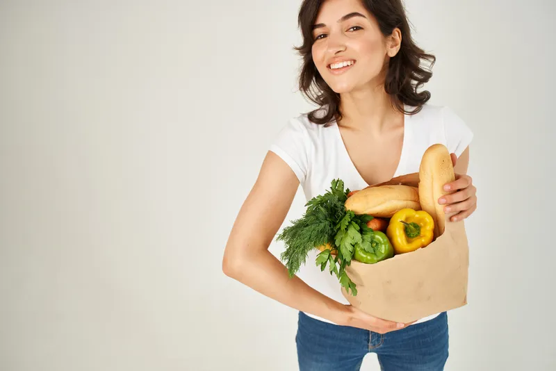 Симпатичная брюнетка в белой футболке держит в руках пакет с продуктами из супермаркета