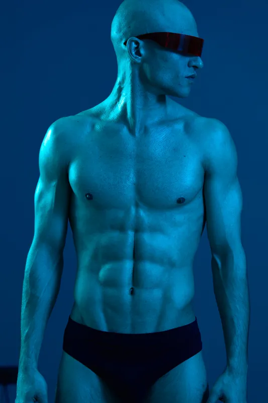 Athletic man posing confidently in blue light, showcasing strong muscles and stylish sunglasses, creating a striking visual impact in a modern setting.