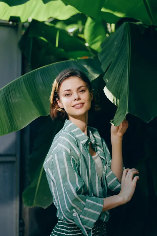 Portrait of a smiling woman in a striped green shirt amid large tropical leaves.