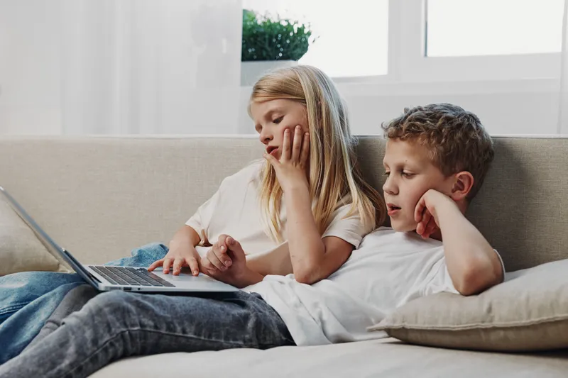 Two children sitting on a sofa using a laptop together in a bright living room. Concept of remote learning, technology, and childhood interaction at home.