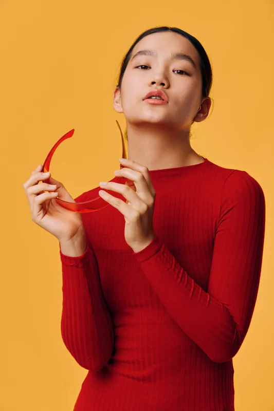 Woman in red long sleeve dress posing against yellow background holding red sunglasses, fashion portrait with confident expression and minimalist style in studio shot.