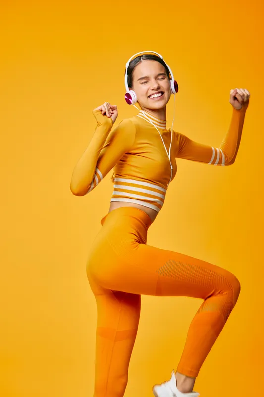 Energetic woman in orange sportswear leaps with headphones, showcasing fitness and sport energy. Dynamic pose, vibrant orange background, upbeat workout mood and athletic lifestyle.