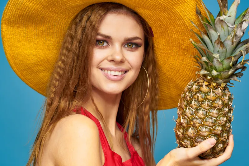 Женщина в жёлтой шляпе держит в руках ананас, у неё вьющиеся волосы, а вокруг экзотические фрукты на голубом фоне