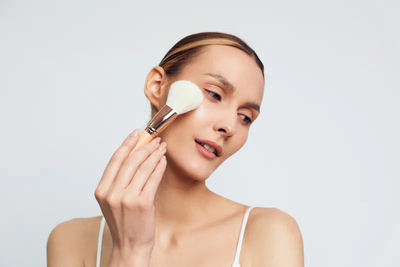 Young woman with fair skin applies makeup using a large brush while looking slightly to the side against a plain light background.