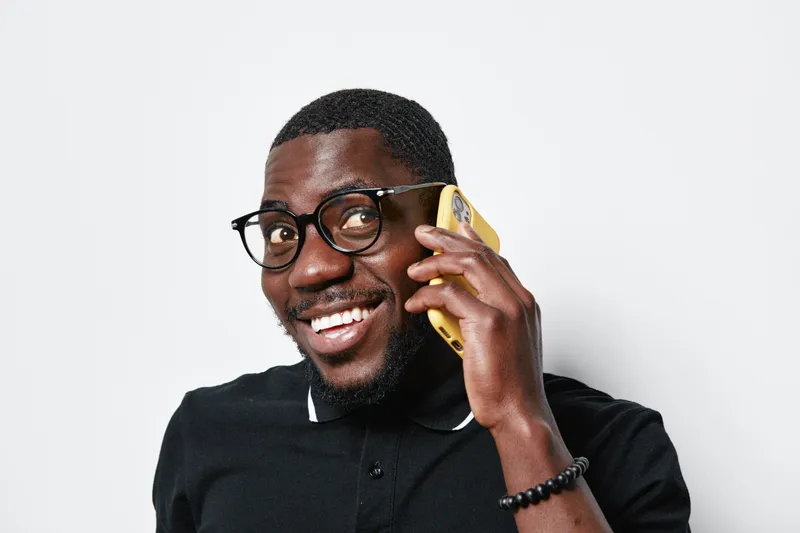 African American man with dark skin wearing glasses and black polo shirt smiling while talking on smartphone against white background. Positive expression and casual style portrait.