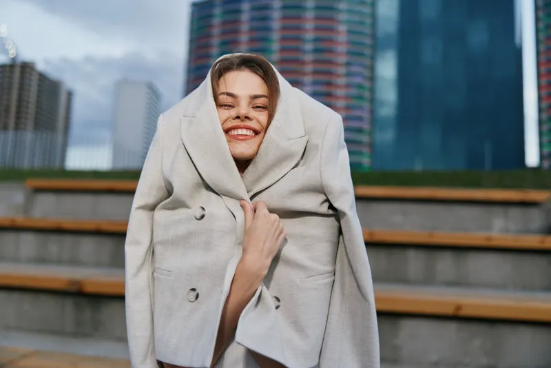 Smiling woman in oversized blazer embracing urban aesthetics with a playful pose, showcasing confidence and creativity in a modern business environment.