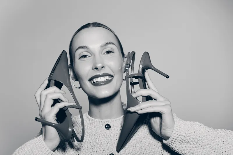 Young white woman with pale skin smiles holding two high heel shoes near her ears. Studio portrait in monochrome. Concept of fashion, happiness, and personal style.