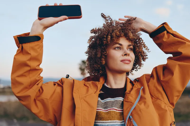 Woman with curly hair in jacket holding smartphone high outdoors, both hands on head, concept woman lifestyle and selfie, communication and technology, bright sky background, natural light, joyful