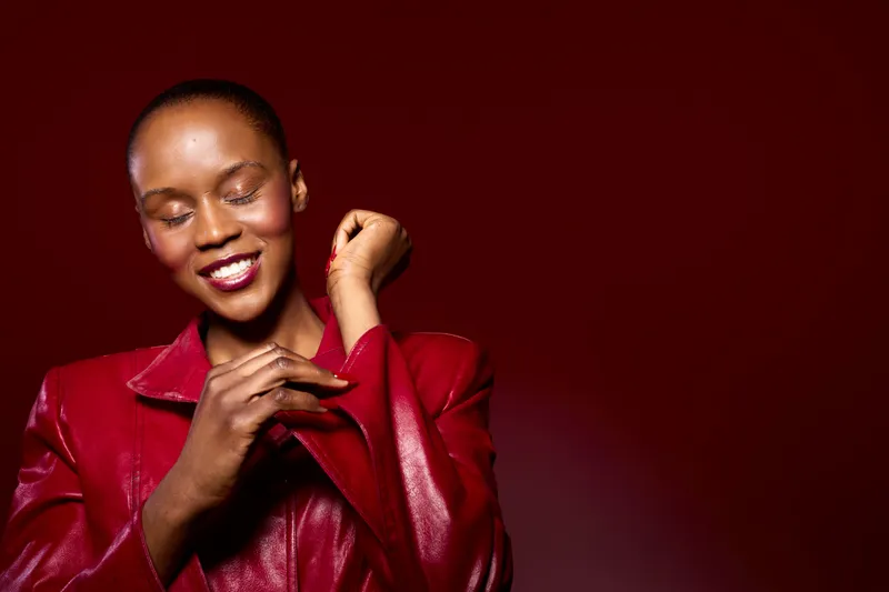 Smiling dark skinned African woman in red leather jacket adjusts sleeve with closed eyes. Studio portrait on dark red background. Concept of fashion, confidence, and beauty with natural skin tone.
