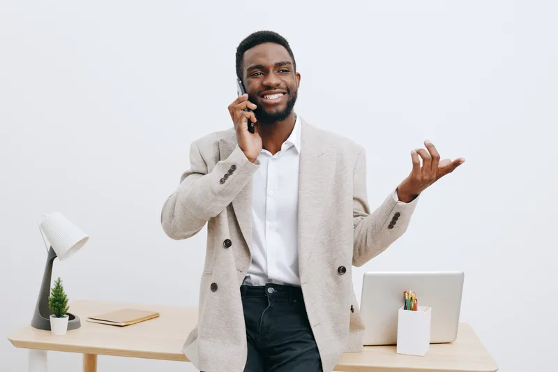 Confident smiling African American man in beige blazer talking on mobile phone while standing at modern office desk, using laptop and stationery, studio shot. Business communication concept