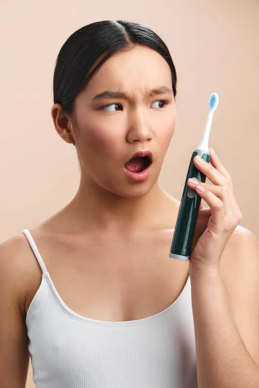 asian woman holds a toothbrush to her ear as if taking a call, with a surprised expression in a simple scene, creating a playful lifestyle moment centered on health and daily routine.