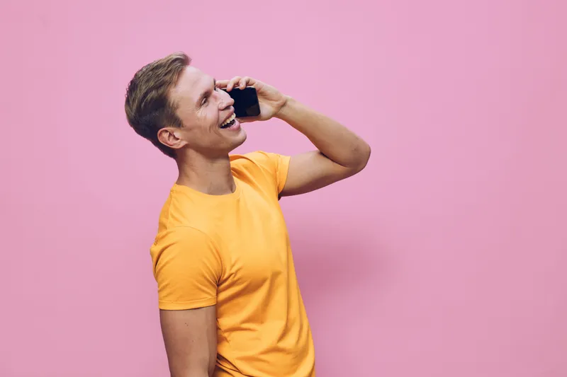 Smiling young man in yellow t-shirt hold smartphone talk phone digital nomad remote work concept against pink background cheerful communication