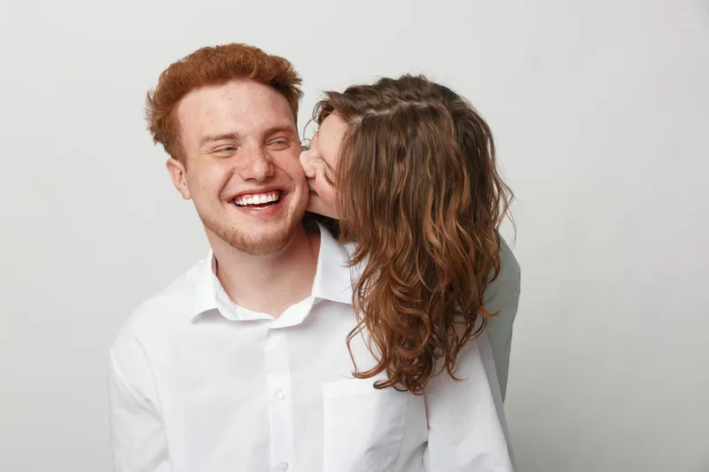 Happy young couple in white shirts share a joyful moment, woman kissing man on cheek. Studio portrait on light background. Concept of love, affection, friendship, and happiness.