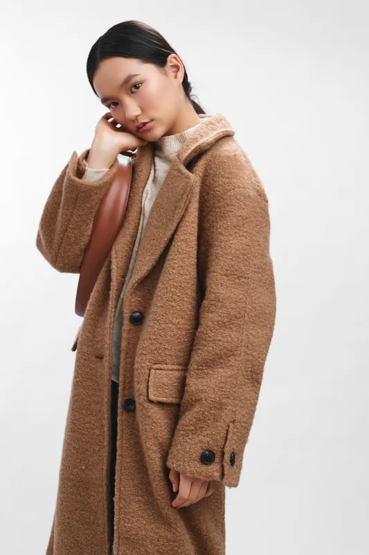 Asian woman wearing a tan wool coat stands thoughtfully with a bag, exuding quiet elegance and confidence in a bright minimalist studio setting, inviting connection and a calm, polished professional