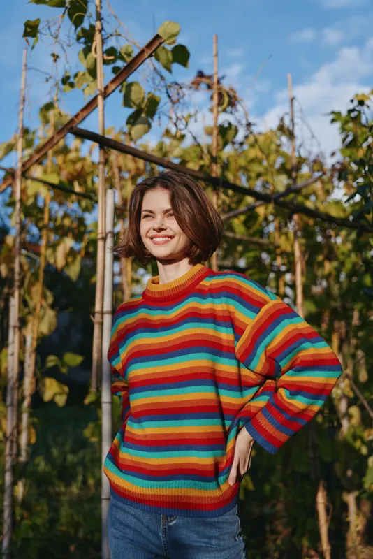 Woman in a colorful striped sweater smiling in a sunlit garden by wooden trellis, short hair, casual outfit with jeans, relaxed pose for lifestyle and fashion.