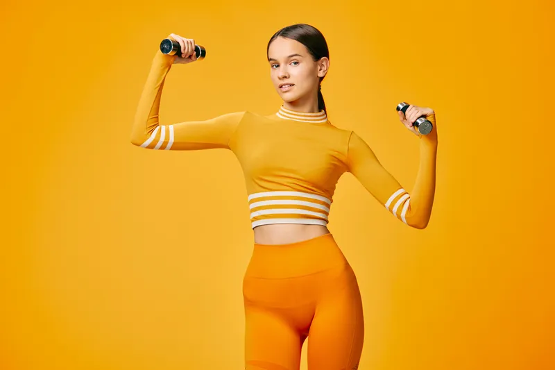 Young fit woman in a matching orange workout set lifting small dumbbells in a bright monochrome studio. Confident pose, toned midriff, active lifestyle and sport concept.