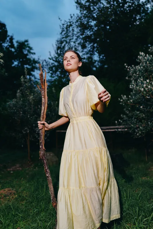A woman in a pale yellow dress stands outdoors in a grassy field, holding a wooden staff, looking upward with a calm expression.