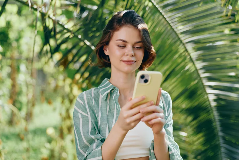 A woman outdoors holds a smartphone, wearing a green striped shirt, focused and calm as she interacts with the device in bright natural light.