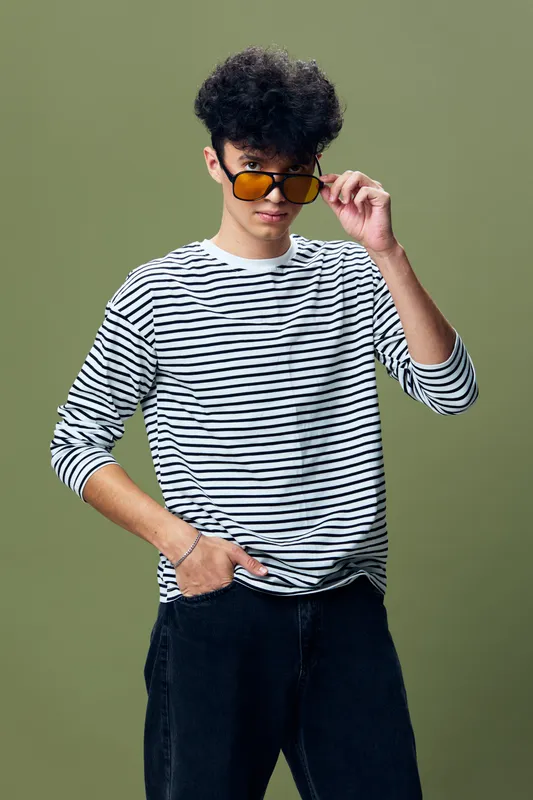 Young man wearing striped long sleeve shirt and yellow sunglasses poses confidently with hand in pocket against green background, showcasing modern casual style.