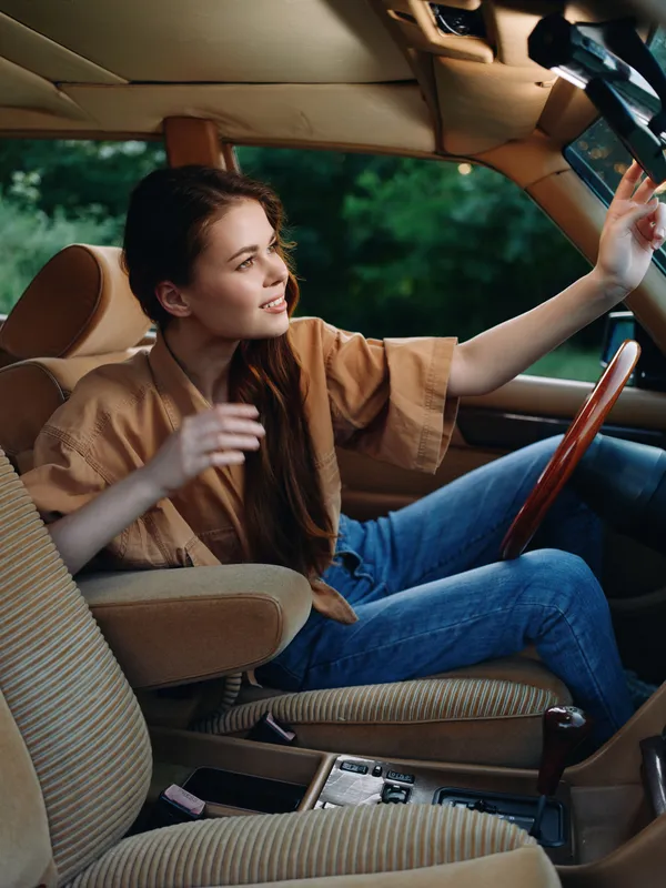 Young woman smiling confidently in a vintage car, wearing a casual outfit, conveying joy and freedom in a lush green setting.