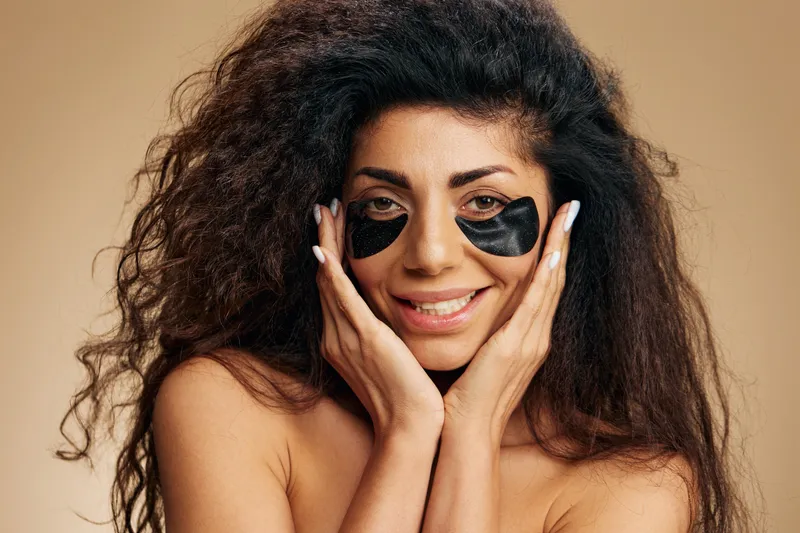 Веселая улыбающаяся красивая латиноамериканка с черными гидрогелевыми патчами под глазами прикасается к идеальной коже и позирует изолированно на пастельном бежевом фоне