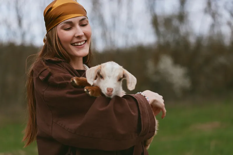 Smiling woman holds a goat kid outdoors in a rustic setting, showing gentle bonding and a lifestyle of simplicity. Warm natural light highlights care, joy, and everyday farm life.