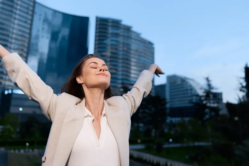 Confident businesswoman with arms outstretched, enjoying life in a modern urban setting, dressed in light professional attire, exuding joy and empowerment.