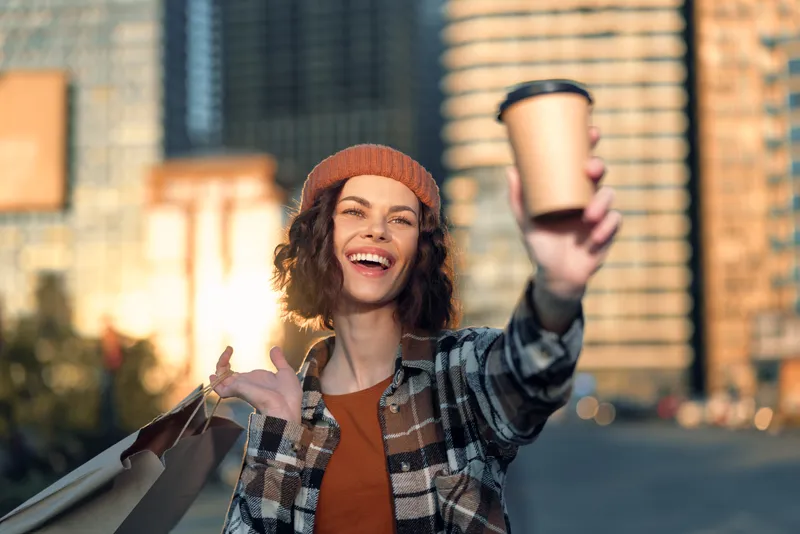 Woman holding coffee and shopping bags smiles in city golden backlight, candid authenticity and mindful living captured in golden hour glow, emotional storytelling portrait of joy.