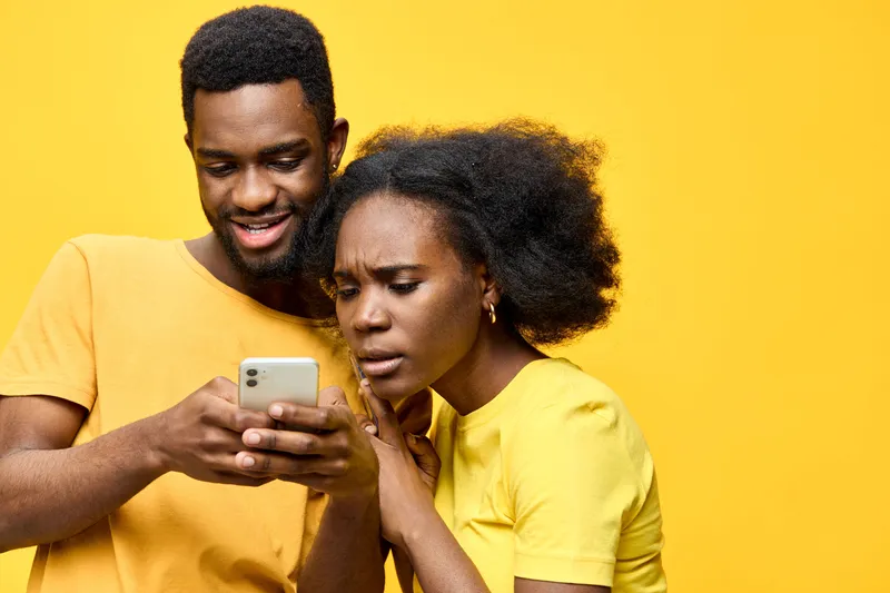 Young African couple interacting with smartphone, expressing curiosity and excitement, vibrant yellow background showcasing a modern and cheerful atmosphere