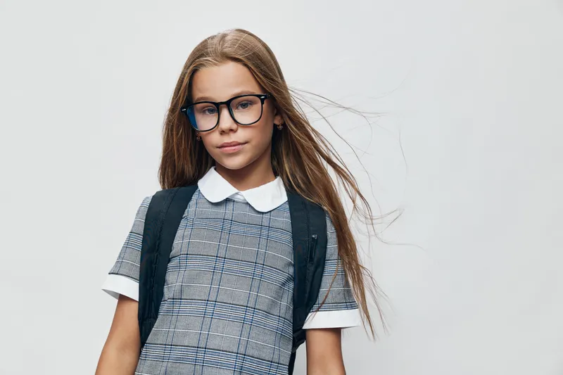 Girl child with long hair and glasses wearing plaid dress and white collar posing with serious face on plain background. Young student with trendy outfit and backpack for school portrait.