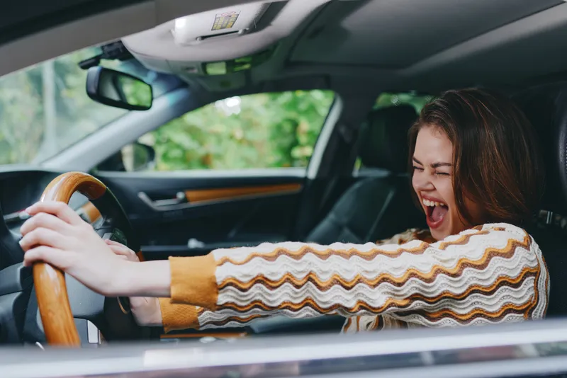 Joyful woman driving a car, gripping the wheel with excitement, laughing aloud as warm interior tones frame a lively, candid moment.