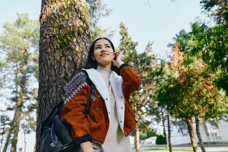 Woman in a warm jacket talks on a phone while strolling outdoors among tall trees, backpack on shoulder, sunlight filtering through leaves, urban park mood, candid moment of everyday connection